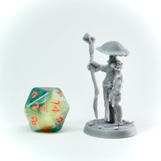Gray figurine of a character with a staff and mushroom-like hat next to a green and orange die on a white background