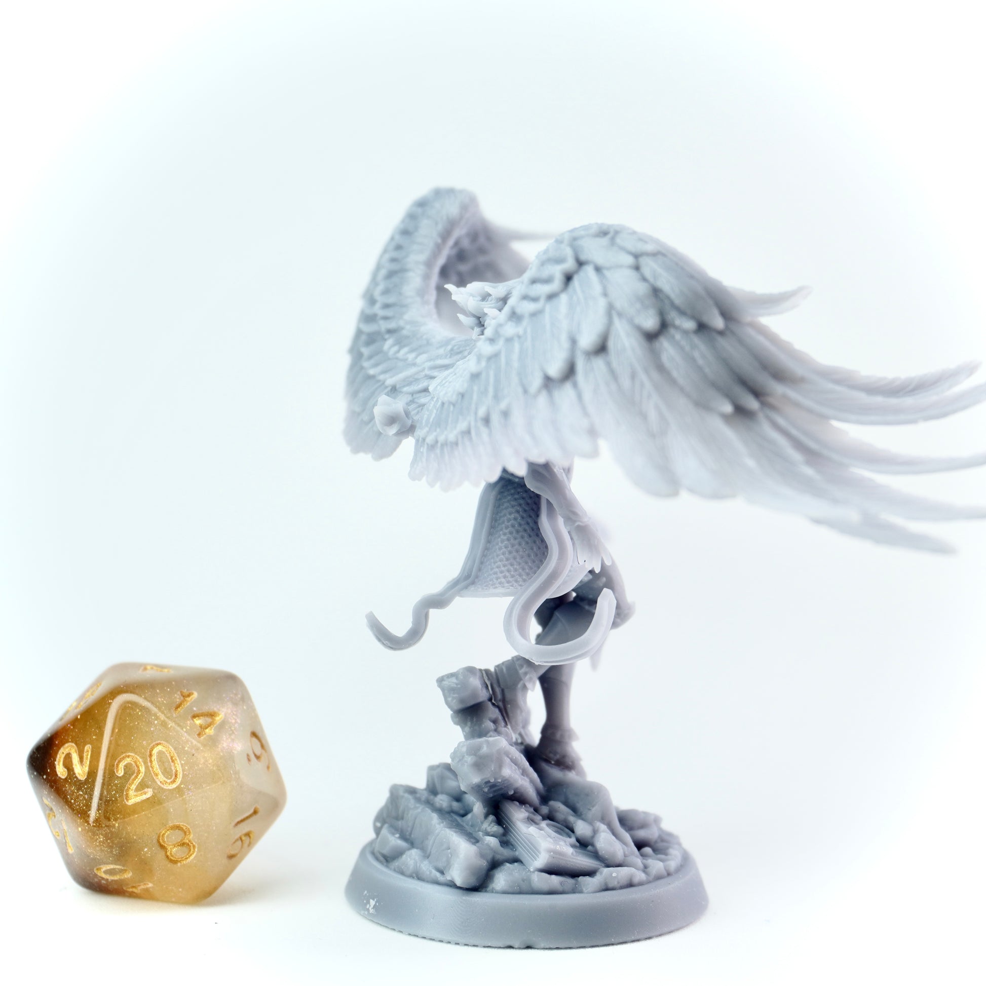 Gray angel figurine with outstretched wings next to a gold twenty-sided die on a white background