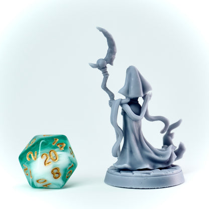 Gray figurine of a evil nun in a hooded robe holding a scythe next to a green polyhedral dice on a white background