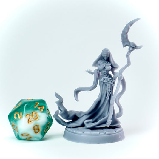 Gray figurine of a evil nun in a hooded robe holding a scythe next to a green polyhedral dice on a white background
