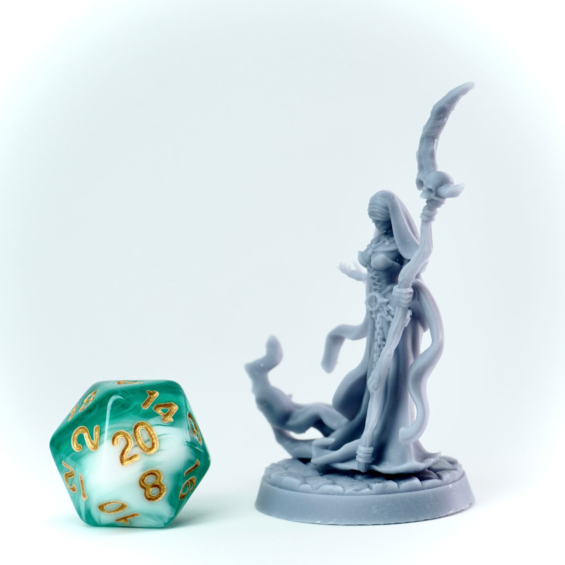 Gray figurine of a evil nun in a hooded robe holding a scythe next to a green polyhedral dice on a white background