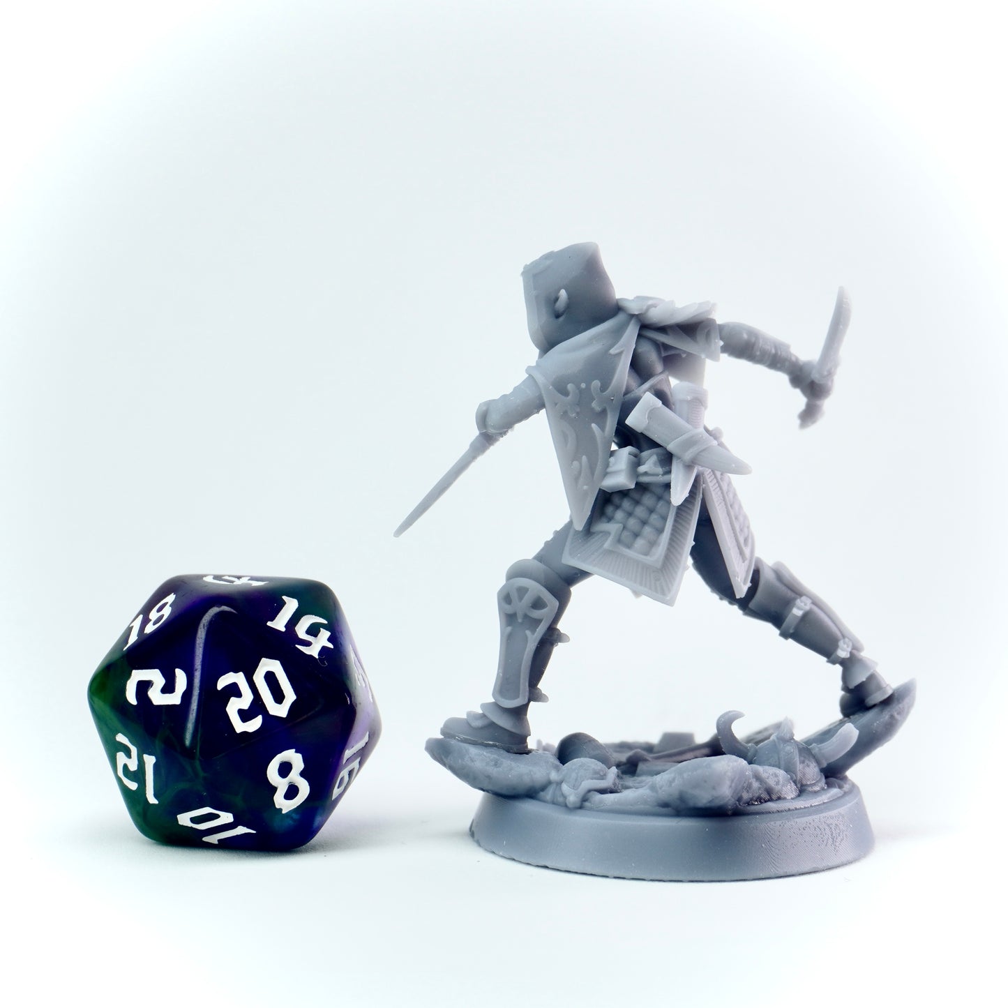Gray miniature figure of a rogue with a daggers, standing on a base, next to a 20-sided die on a white background.