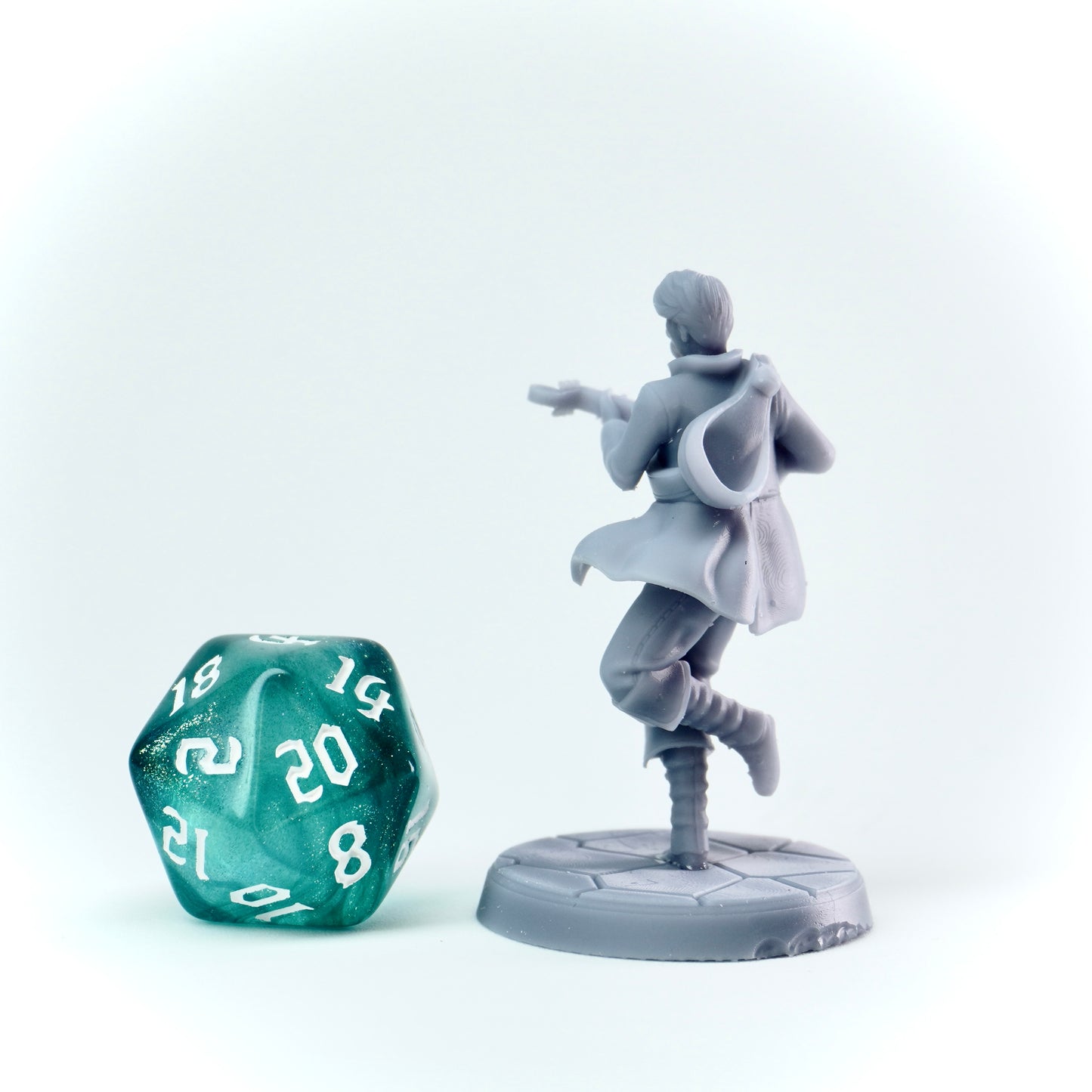 Gray miniature figure of a character in a cloak next to a green polyhedral dice on a white background