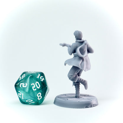 Gray miniature figure of a character in a cloak next to a green polyhedral dice on a white background