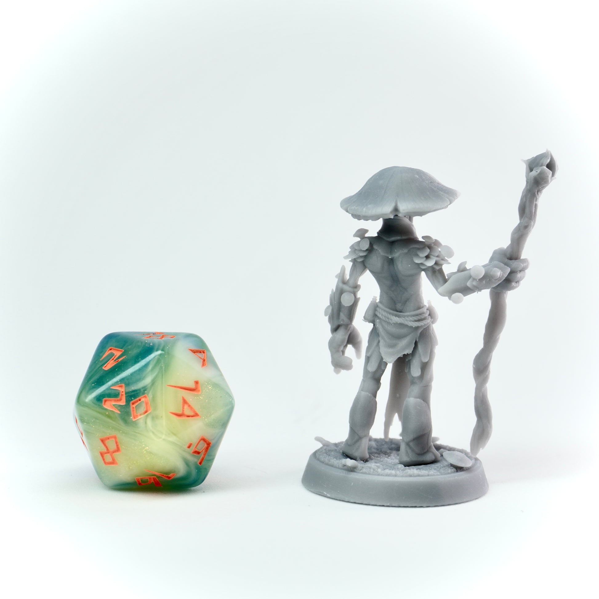Gray miniature figure with a staff next to a green and yellow polyhedral dice on a white background