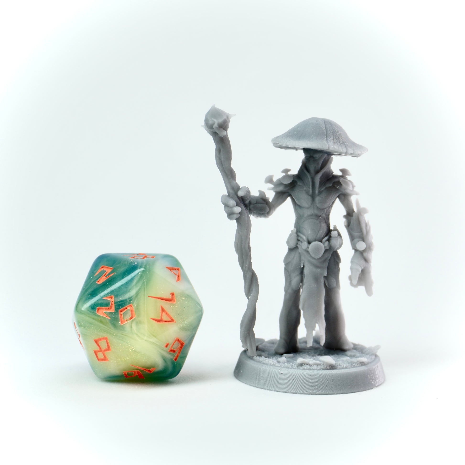 Gray fantasy figurine holding a staff next to a green and orange polyhedral dice on a white background