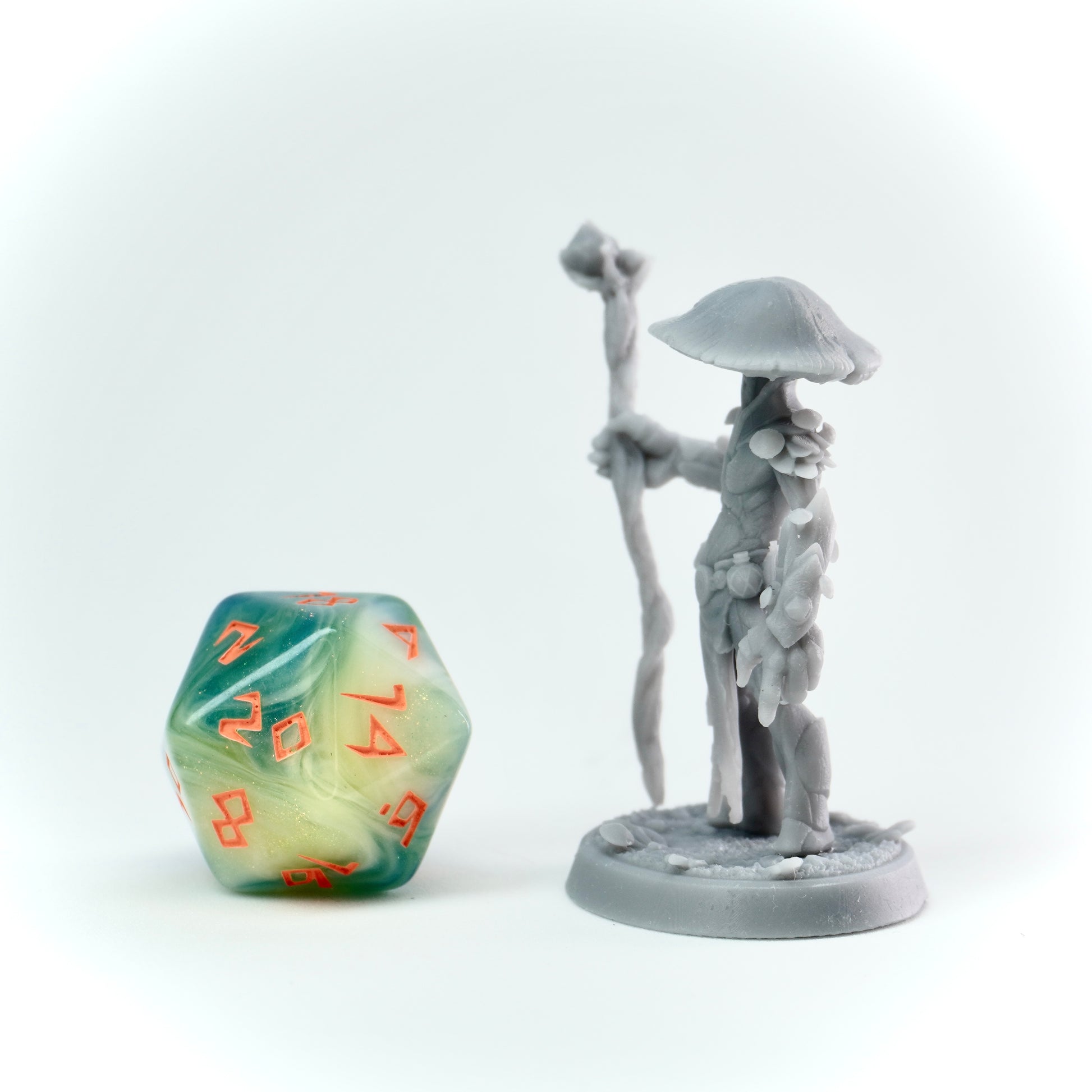Gray figurine of a character with a staff and mushroom-like hat next to a green and orange die on a white background