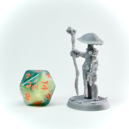 Gray figurine of a character with a staff and mushroom-like hat next to a green and orange die on a white background
