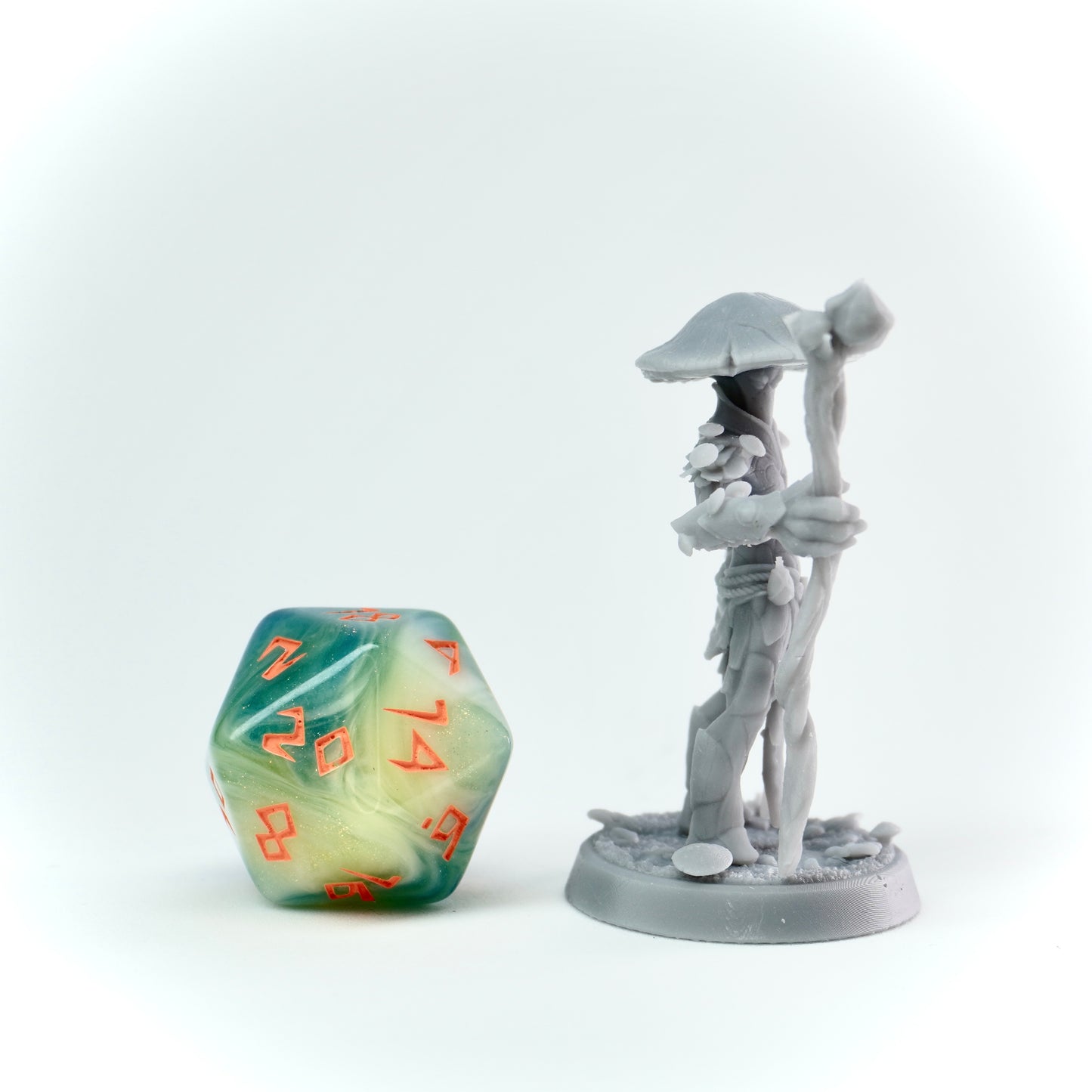 Gray miniature figure of a muconid with a staff next to a green polyhedral dice on a white background
