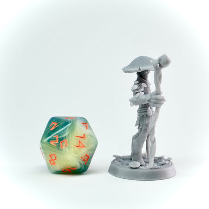 Gray miniature figure of a muconid with a staff next to a green polyhedral dice on a white background