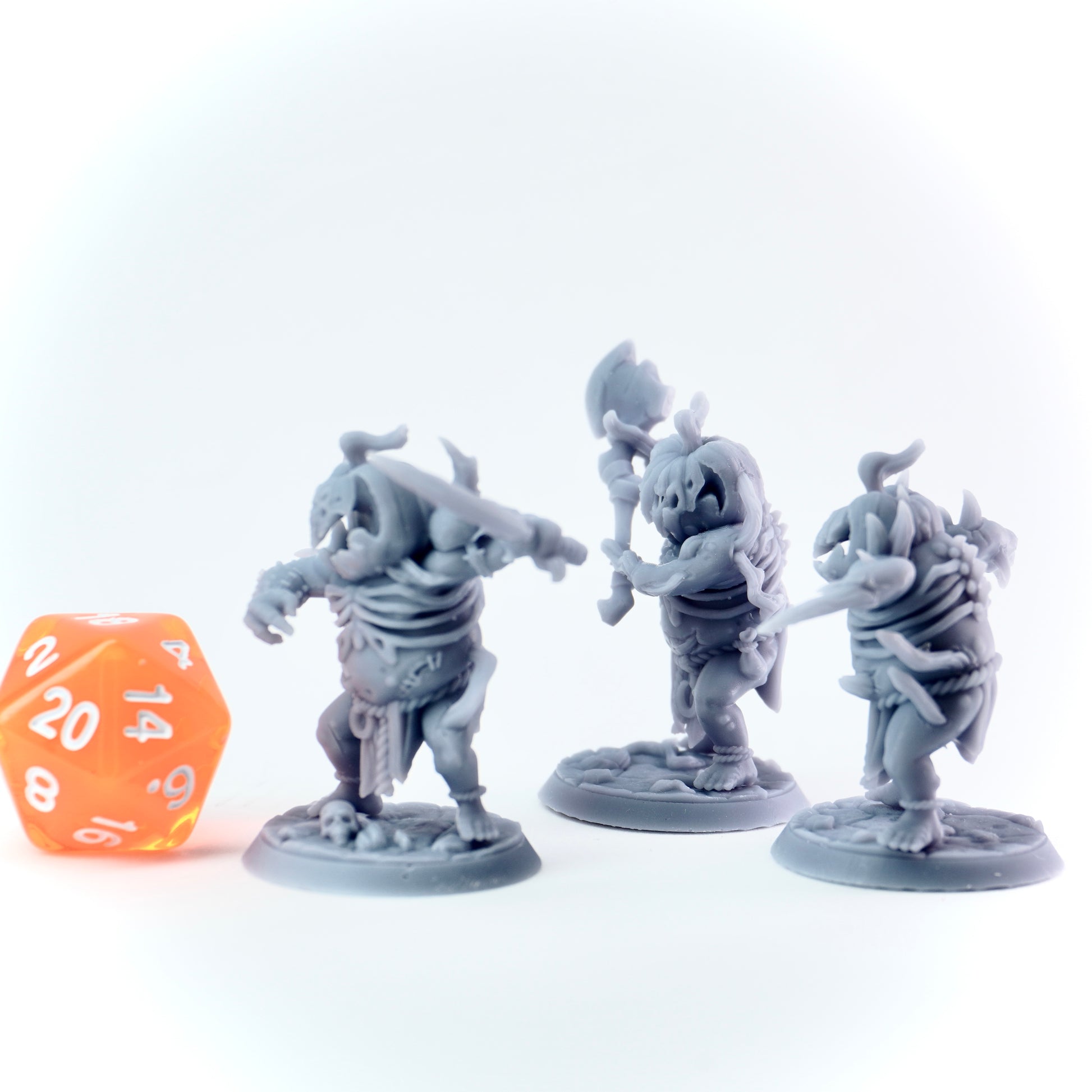 Three gray miniature figures with weapons on a white background, accompanied by an orange die.