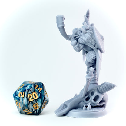 Gray fantasy frog figurine with a large weapon and dead animal skull next to a blue and gold dice on a white background