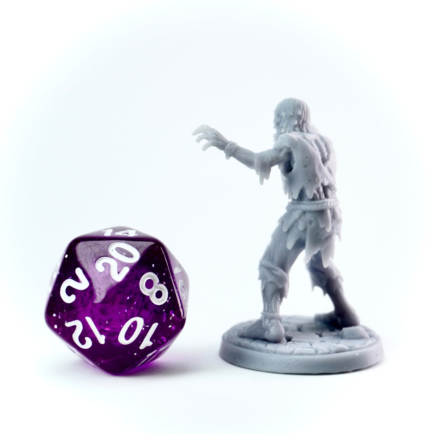 Gray zombie miniature next to a purple polyhedral dice on a white background