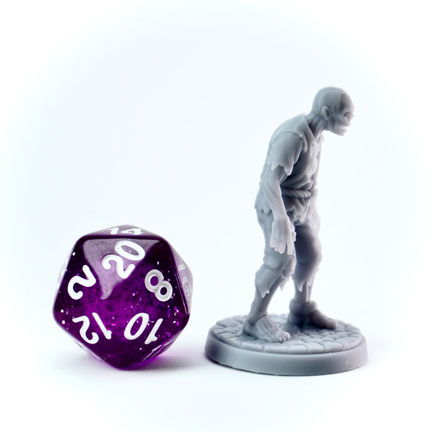 Gray zombie miniature of a humanoid standing next to a purple polyhedral dice on a white background