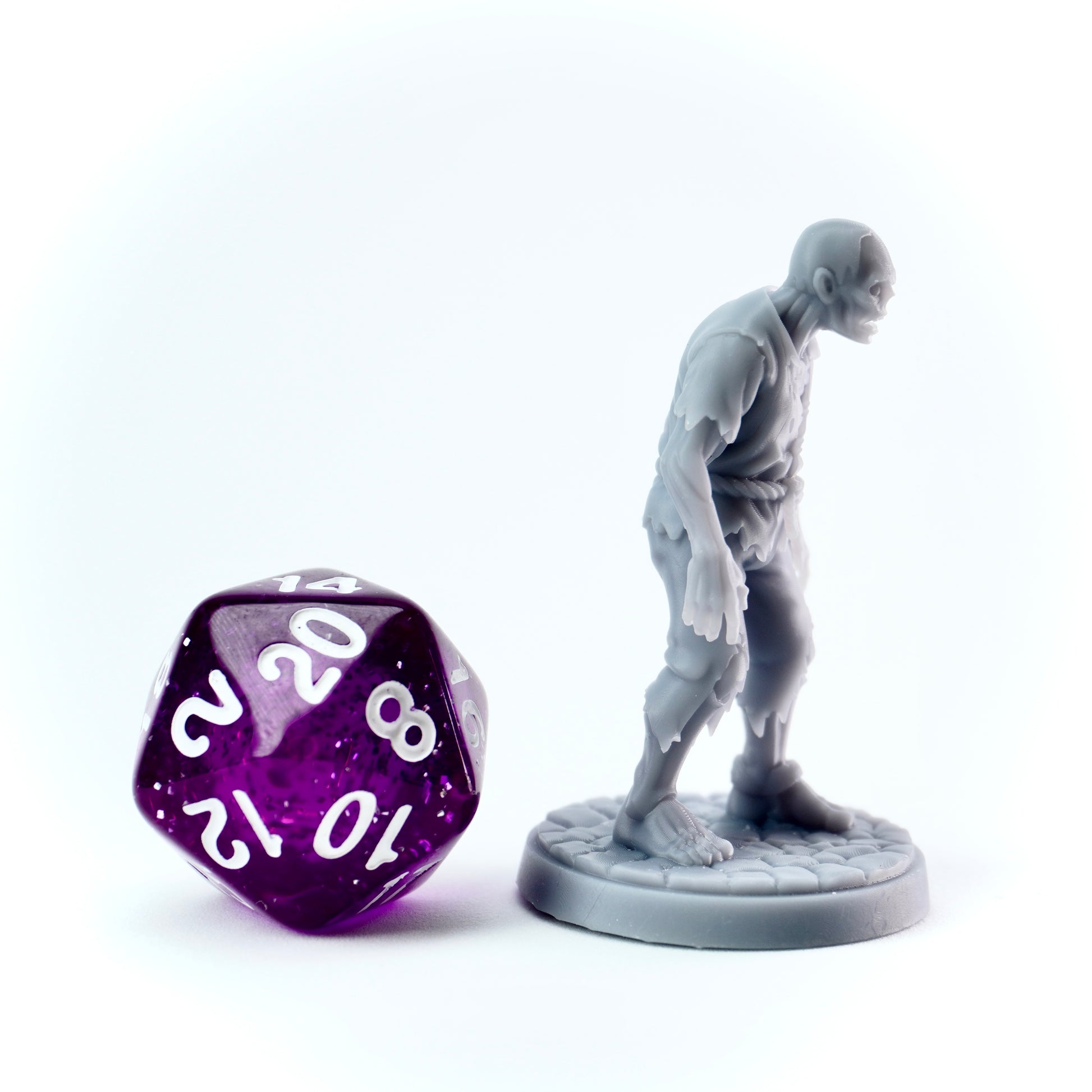Gray zombie miniature of a humanoid standing next to a purple polyhedral dice on a white background