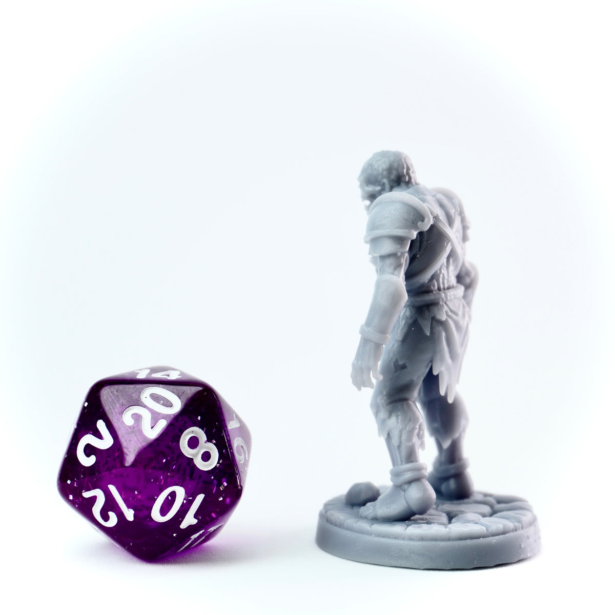 Gray miniature figurine of a warrior next to a purple polyhedral dice on a white background