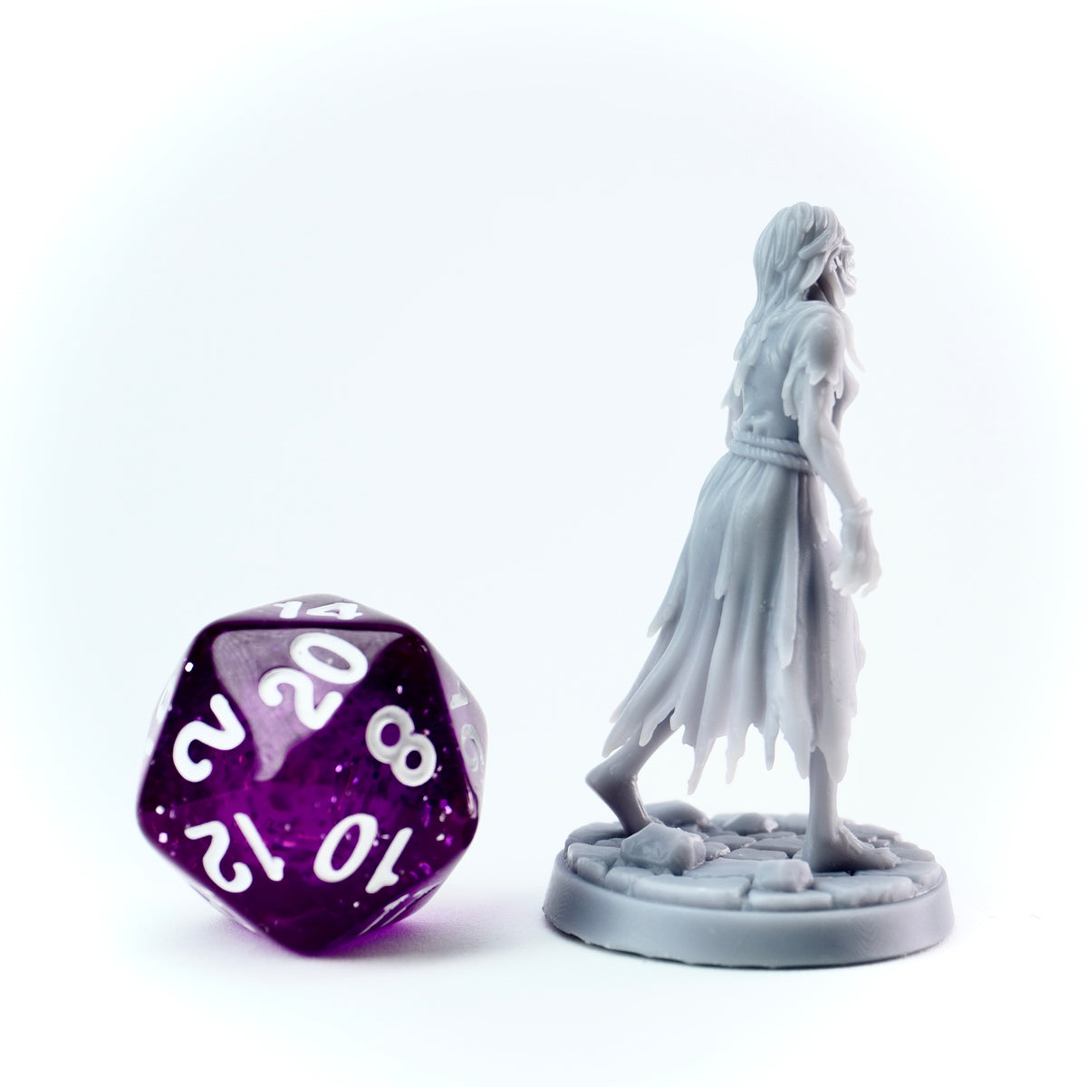 Gray figurine of a hooded figure next to a purple polyhedral dice on a white background